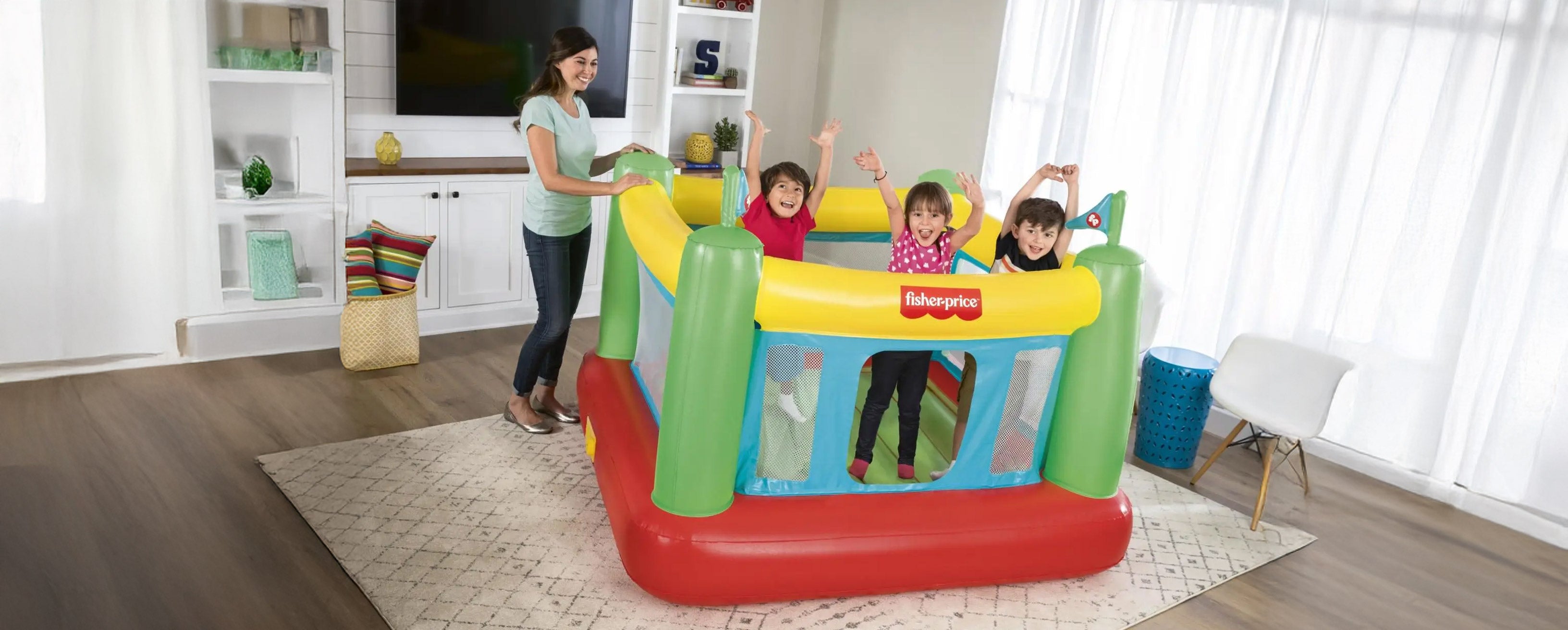 Three smiling kids jumping on a Fisher-Price Bouncesational indoor bounce house with a built-in pump, with a parent nearby.
