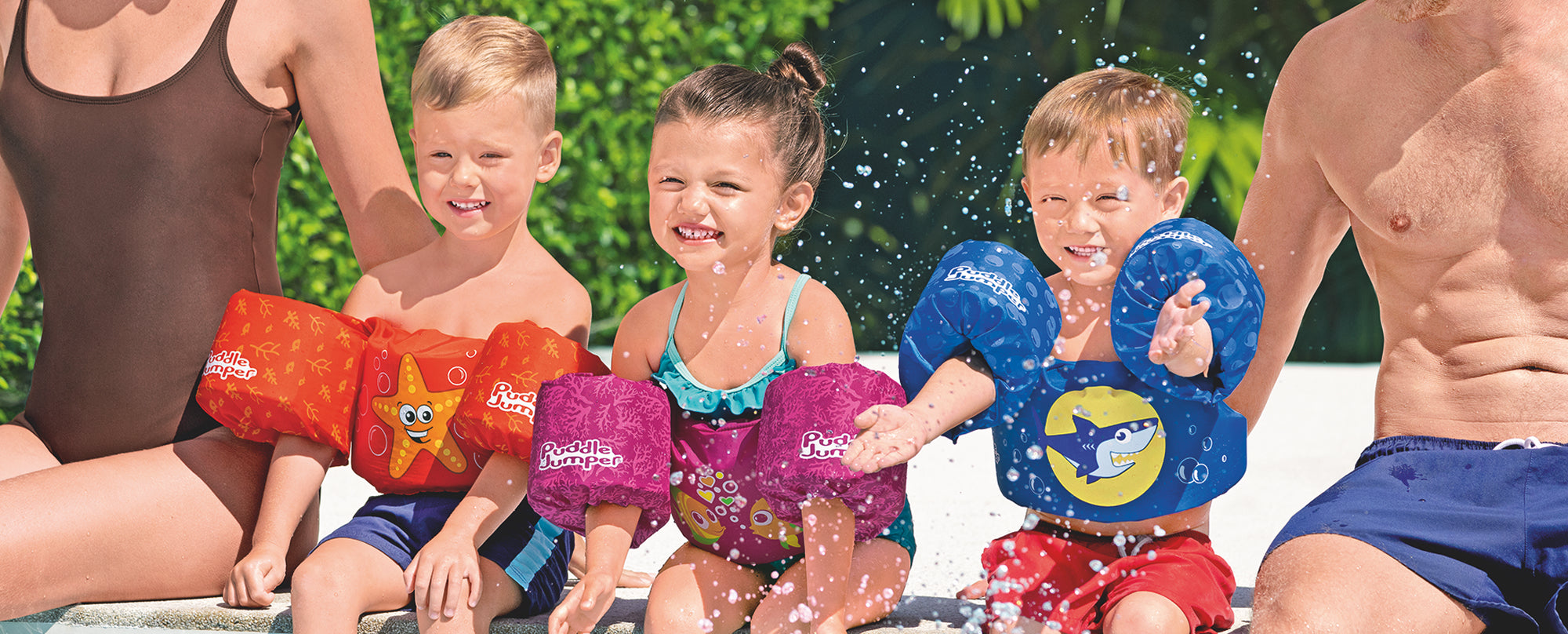 Three young children are wearing child life vests and are sitting on the edge of a pool with two adults.